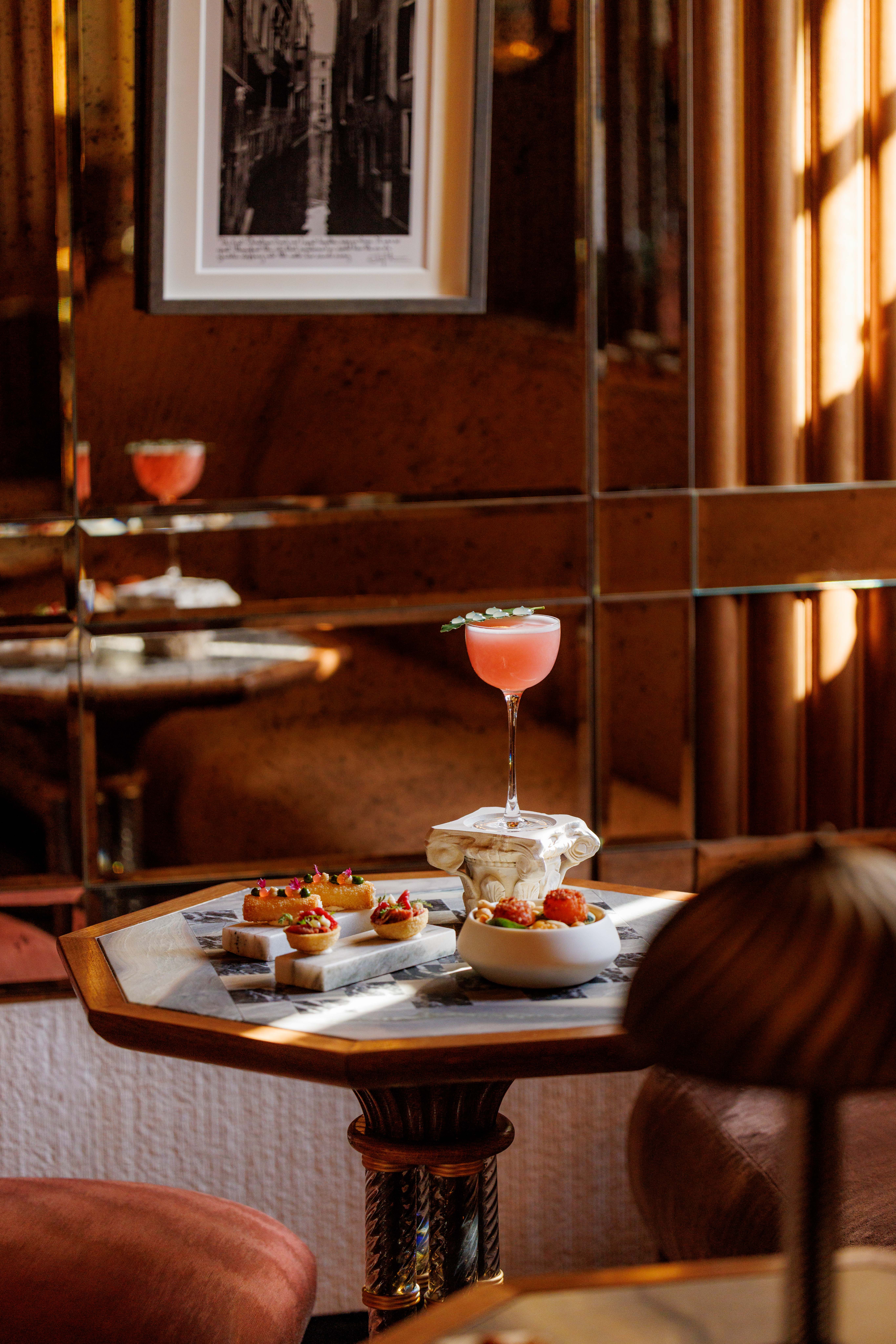 Pink coupe cocktail and a plate of petits fours on a table in an elegant hotel bar at Orient Express Venezia, Italy