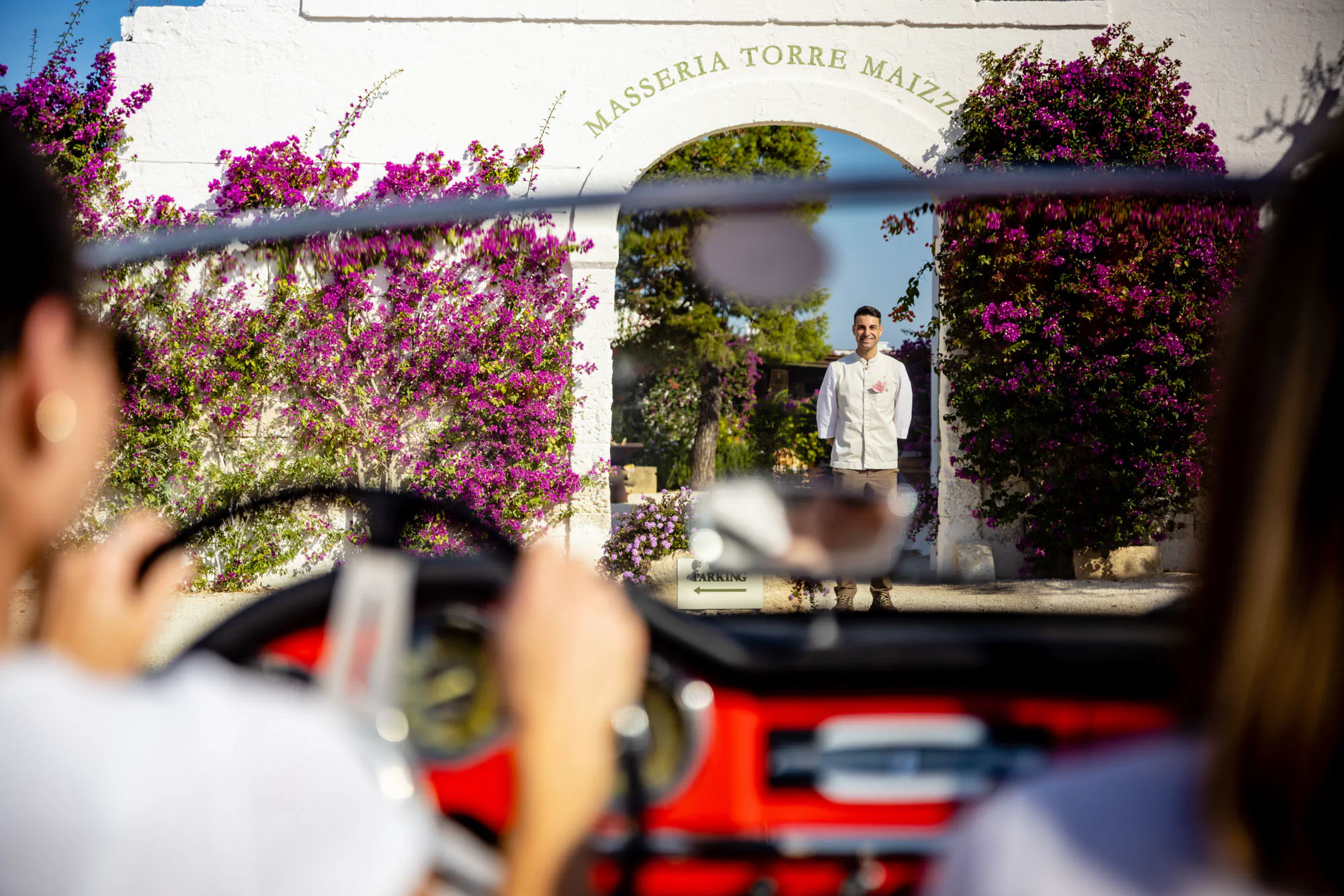 Red convertible arriving at a villa entrance framed by bougainvillea, a host greeting