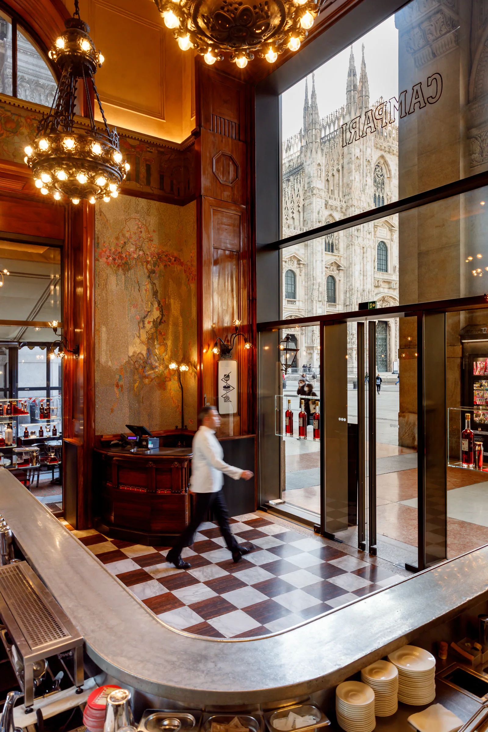 Opulent hotel lobby with chandeliers and a checkered floor, the Milan Duomo visible outside