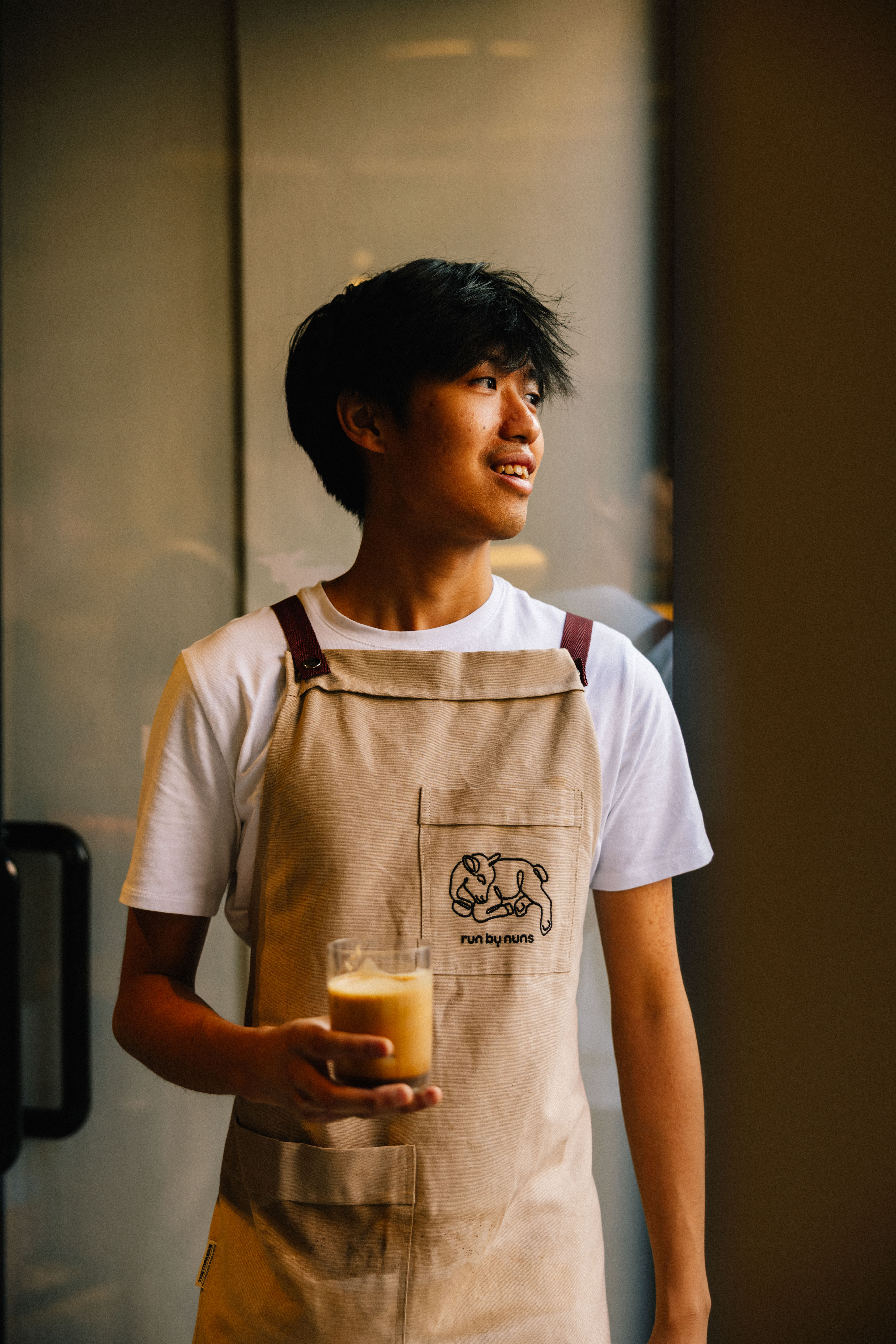 Young person in an apron holding an iced drink by a window